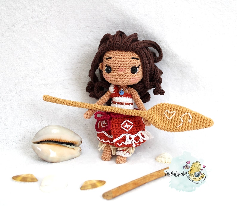 May include: A crocheted doll of a young girl with brown hair, wearing a red and white dress and holding a wooden paddle. The doll is standing on a white surface with seashells.