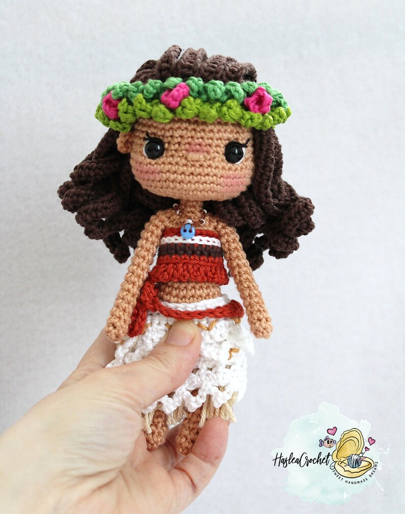 May include: Crocheted doll with brown hair, wearing a green and pink flower crown, a red and white top, and a white skirt. The doll is holding a blue and white necklace with a small fish charm.