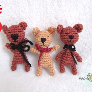 Puede incluir: Tres osos de peluche tejidos a crochet, uno marrón, uno beige y uno marrón con un lazo negro. Los osos están todos de pie sobre una superficie blanca.