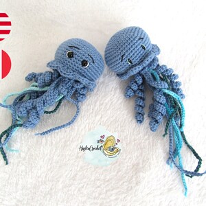Puede incluir: Dos juguetes de medusa de crochet azul con tentáculos verdes y azules. Las medusas tienen ojos negros y una expresión sonriente. Los juguetes están sobre una superficie blanca y esponjosa.