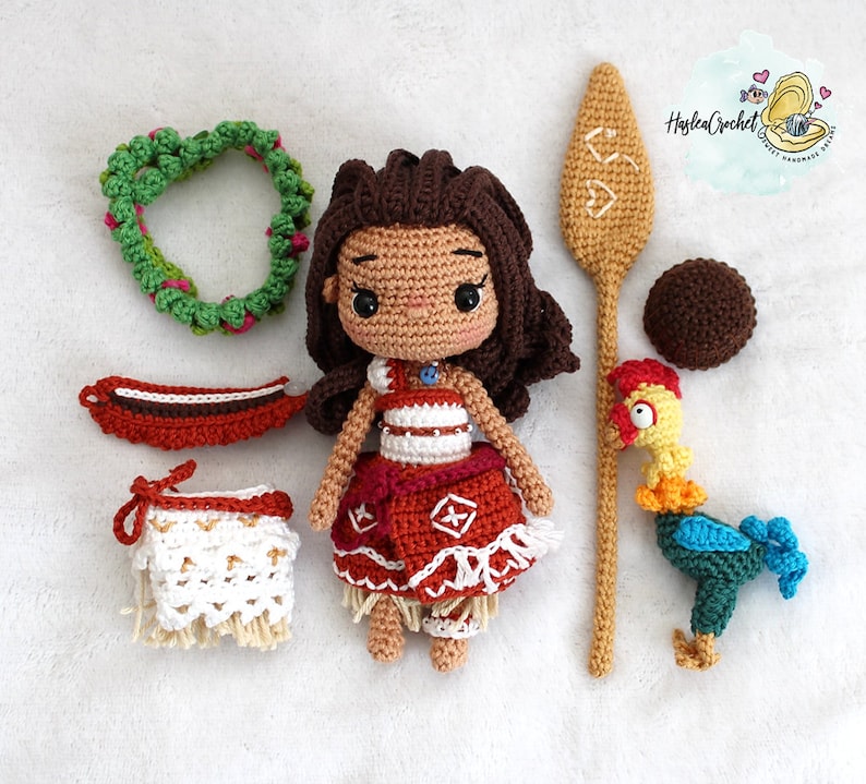 May include: Crocheted amigurumi doll of Moana from Disney's Moana, wearing a red and white dress, a lei, and a blue necklace. The doll is surrounded by other crocheted items, including a rooster, a paddle, and a flower lei.