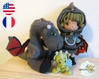 Peut inclure: Une poupée au crochet portant une cape à capuche verte et tenant un petit dragon vert. Un plus grand dragon gris aux ailes rouges est assis à côté d'eux.