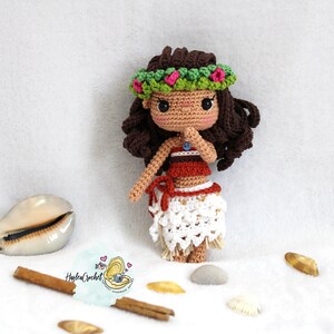 May include: Crocheted doll with brown hair wearing a green flower crown, red and brown top, and a white skirt. The doll is standing on a white surface with seashells and cinnamon sticks.