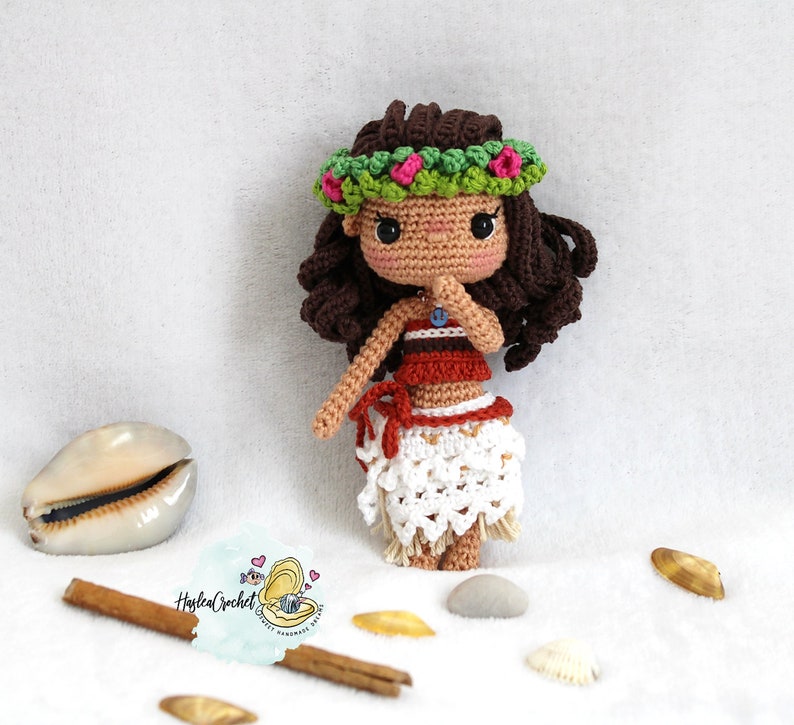 May include: Crocheted doll with brown hair wearing a green flower crown, red and brown top, and a white skirt. The doll is standing on a white surface with seashells and cinnamon sticks.
