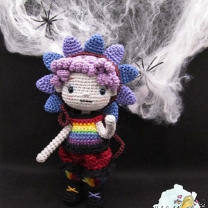 May include: A crocheted doll with a rainbow shirt and a purple flower on its head. The doll is standing in front of a white spider web.