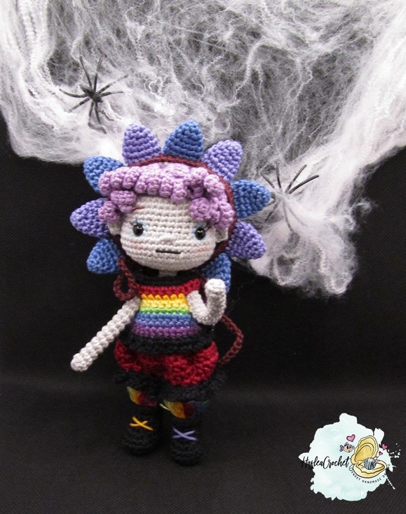 May include: A crocheted doll with a rainbow shirt and a purple flower on its head. The doll is standing in front of a white spider web.