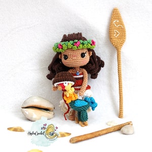 May include: Crocheted amigurumi doll of Moana from Disney's Moana, wearing a flower lei and holding a crocheted rooster. The doll is standing on a wooden surface with a brown stick and a seashell.