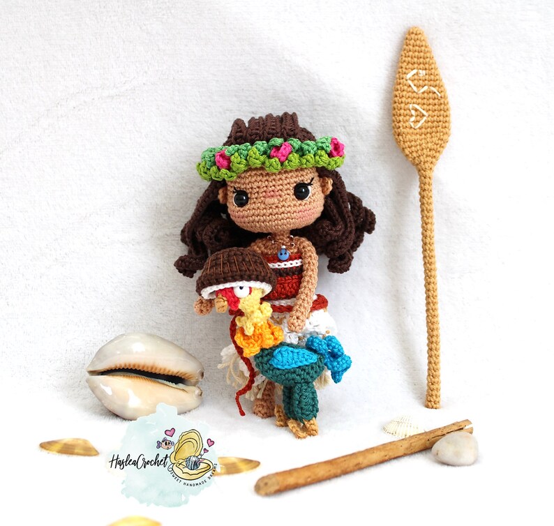 May include: Crocheted amigurumi doll of Moana from Disney's Moana, wearing a flower lei and holding a crocheted rooster. The doll is standing on a wooden surface with a brown stick and a seashell.