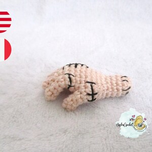 Puede incluir: Una mano de crochet con puntadas negras, que se asemeja a una herida cosida. La mano es rosa y está sobre una superficie blanca y esponjosa.