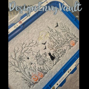 May include: A light gray fabric held in a blue embroidery hoop featuring a black line art design of a witch reading a book, a ghost, a black cat, a haunted house, bare trees, bats, a spider web, and orange pumpkins with embroidered details.