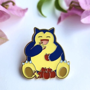 Op de afbeelding: Een gouden emaille pin met een cartoon Snorlax Pokémon die zit en rode appels eet. De Pokémon is geel met blauwe accenten en een grote glimlach.