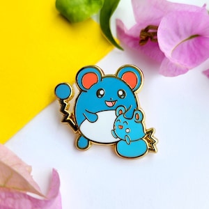 May include: Gold-coloured enamel pin featuring a blue and white Pokemon character with a smaller blue Pokemon character in its arms. The larger Pokemon has red ears and a yellow lightning bolt tail.