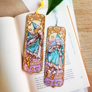 May include: Two illustrated bookmarks with anime-style characters in blue and white robes and hats. The bookmarks have gold and purple borders and yellow tassels. The bookmarks are placed on an open book, with text visible.