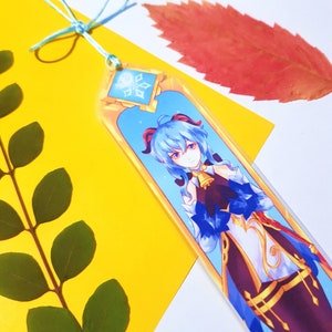 May include: A blue and gold bookmark featuring an anime character with blue hair and a snowflake design. The bookmark has a blue ribbon attached.