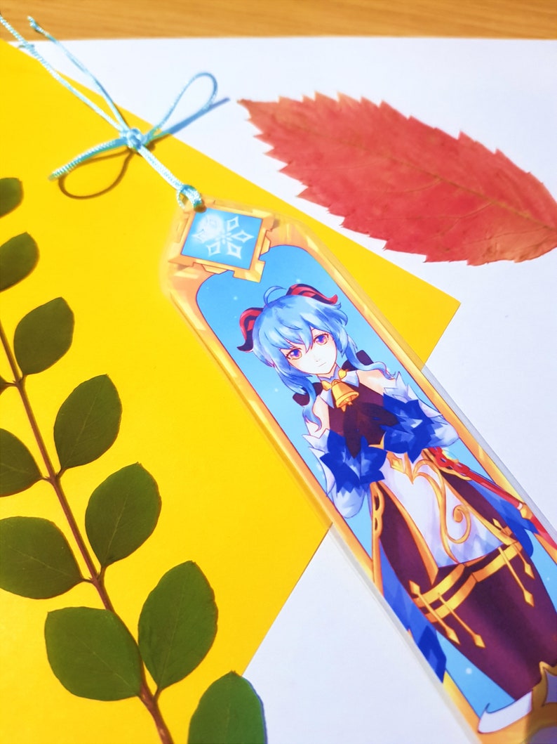May include: A blue and gold bookmark featuring an anime character with blue hair and a snowflake design. The bookmark has a blue ribbon attached.