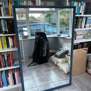 May include: A large, rectangular mirror with a dark blue frame. The frame is decorated with a row of small, round beads. The mirror is reflecting a room with bookshelves and a black bag.