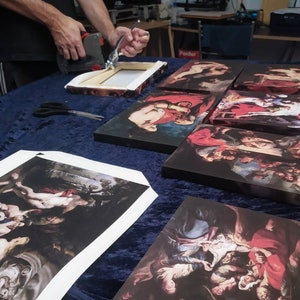 May include: A person is stapling a canvas print to a wooden frame. The print is a reproduction of a painting with a group of people in a dark setting. The print is on a table with other canvas prints.