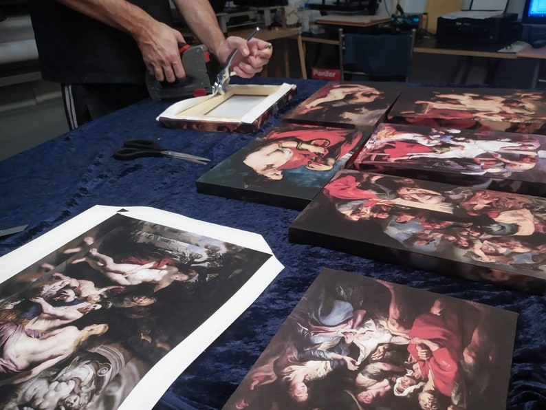 May include: A person is stapling a canvas print to a wooden frame. The print is a reproduction of a painting with a group of people in a dark setting. The print is on a table with other canvas prints.