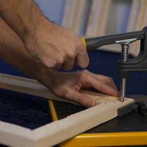 May include: A person uses a black and yellow clamp to secure a wooden frame. The clamp has a metal screw and a handle with the text "FM2010" printed on it.
