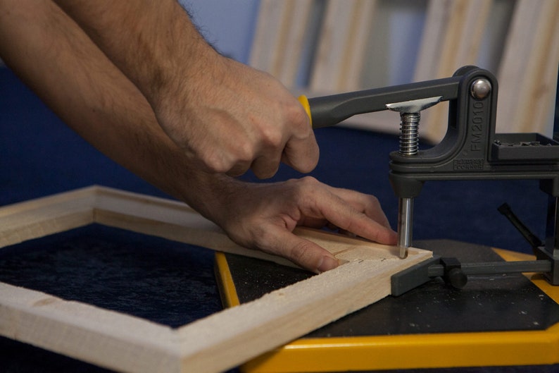 May include: A person uses a black and yellow clamp to secure a wooden frame. The clamp has a metal screw and a handle with the text "FM2010" printed on it.