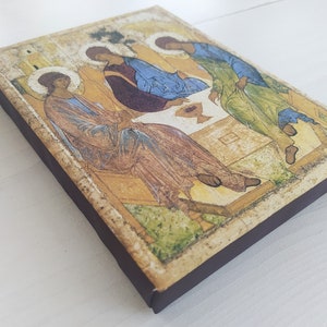 May include: A wooden icon depicting the Holy Trinity, with three figures in robes, sitting around a table with a chalice. The icon has a distressed, aged look and is framed with a dark brown border.