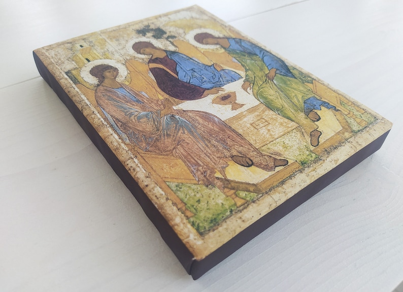 May include: A wooden icon depicting the Holy Trinity, with three figures in robes, sitting around a table with a chalice. The icon has a distressed, aged look and is framed with a dark brown border.