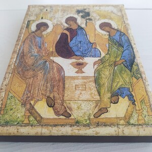 May include: A vintage-style religious icon depicting three figures in robes, seated around a table with a chalice. The figures have halos and are painted in muted tones of brown, blue, and green. The background is a faded gold.