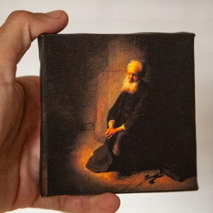 May include: A painting of an older man with a white beard, wearing a dark robe, sitting with his hands clasped in front of him. The painting is set against a dark background with a warm light source illuminating the man.
