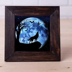 May include: A framed artwork featuring a wolf howling at a blue moon with bats flying around it. The artwork is set against a dark blue starry sky.