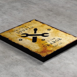 Puede incluir: Una impresión en lienzo amarillenta y desgastada con texto negro que dice "No puedo arreglar la estupidez... pero puedo cobrar por ello." Una llave inglesa y un destornillador negros se cruzan en el centro de la impresión.