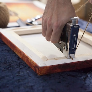 May include: A person using a staple gun to attach twine to a wooden canvas frame. The frame is covered in white canvas and has a red and orange border.