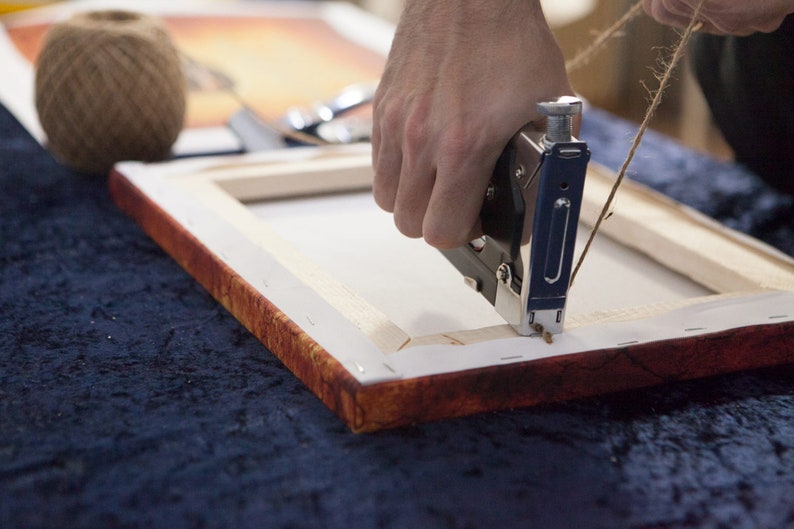 May include: A person using a staple gun to attach twine to a wooden canvas frame. The frame is covered in white canvas and has a red and orange border.