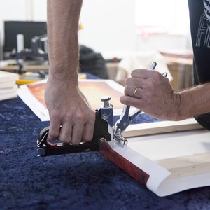 May include: A person using a hand-held stapler to attach a canvas print to a wooden frame. The canvas print has a red and orange abstract design.