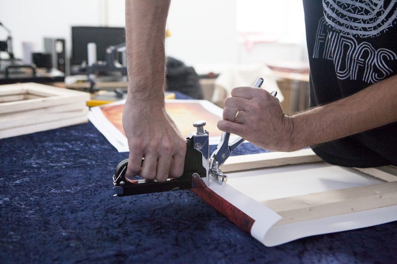May include: A person using a hand-held stapler to attach a canvas print to a wooden frame. The canvas print has a red and orange abstract design.