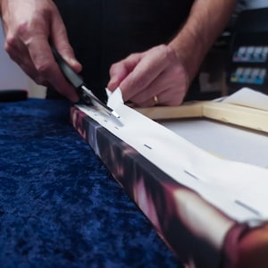 May include: A person is using a tool to attach a white canvas to a wooden frame. The canvas is being stretched tightly over the frame. The frame is on a blue surface.