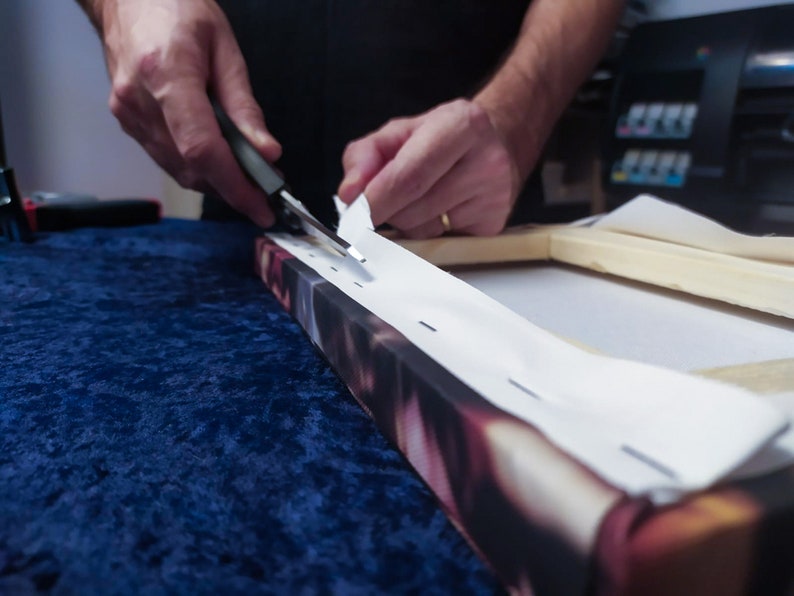 May include: A person is using a tool to attach a white canvas to a wooden frame. The canvas is being stretched tightly over the frame. The frame is on a blue surface.