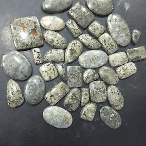 Puede incluir: Una colección de piedras preciosas de pirita en varias formas, incluyendo óvalos y rectángulos. Las piedras tienen un color gris metálico con una apariencia moteada y texturizada. Las piedras preciosas están dispuestas sobre una superficie oscura.