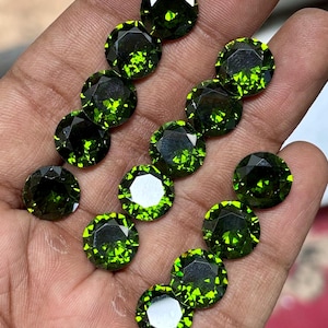 May include: A collection of faceted, round gemstones in various sizes. The gems are a deep, vibrant green with a brilliant, reflective quality. The facets catch the light, creating a sparkling effect. The gems are displayed against a neutral background.
