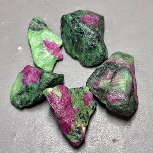 May include: Five rough-cut ruby in zoisite stones. The stones display a mix of deep green and pinkish-red hues. The raw gemstones are irregularly shaped and have a natural, unpolished appearance.