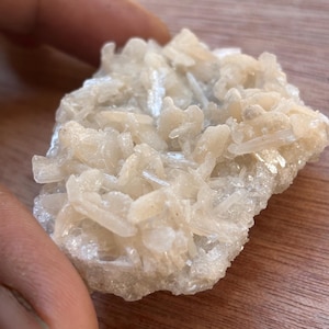 May include: A close-up of a raw, natural crystal cluster. The crystal formation is a pale, creamy white color with a rough, textured surface. The crystals are clustered together, creating a layered appearance. The overall shape is irregular and organic.