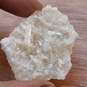May include: A close-up of a light-colored mineral specimen. The mineral has a crystalline structure with numerous small, elongated crystals clustered together. The color is a pale, translucent off-white, with a slightly rough texture. The specimen is held by a person's fingers.