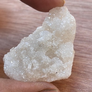 May include: A rough, off-white crystal cluster with a sparkling, granular texture. The natural mineral formation is held by a hand, showcasing its irregular shape and subtle variations in color. The background is a warm-toned wooden surface.