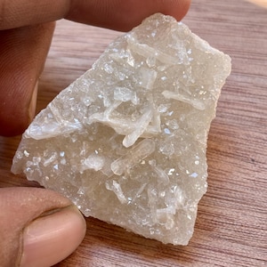 May include: A light beige mineral specimen with crystalline structures. The rough-hewn piece has a textured surface and a mix of small and elongated crystals. The mineral is held in a hand, with a wooden surface in the background.