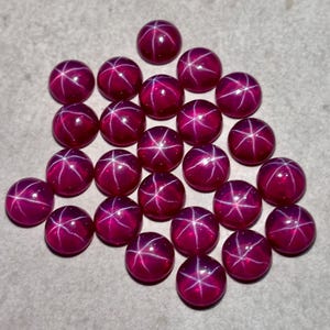 May include: A collection of round, polished, ruby-coloured gemstones. Each gem displays a six-rayed star pattern, with white lines radiating from the centre. The gems are arranged closely together, creating a visually appealing display.