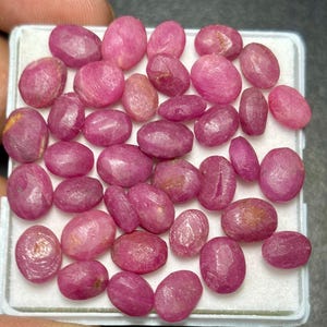 May include: A collection of numerous oval-shaped, translucent pinkish-red rubies arranged on a white foam display pad. Each gemstone exhibits a smooth, polished surface with natural inclusions and varying shades of deep magenta and light pink.