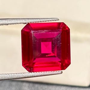 May include: A vibrant, deep red gemstone with a square step-cut facet pattern. The stone features sharp, clean edges and a highly reflective, transparent surface that displays a consistent, saturated crimson hue throughout its entire structure.