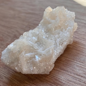 May include: A raw, off-white crystal cluster with a rough, textured surface. The mineral formation has a crystalline structure with multiple points and facets. The natural stone is displayed on a wooden surface.