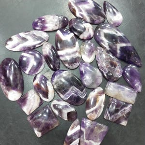 May include: A collection of 20 amethyst cabochons, featuring a variety of shapes and sizes. The cabochons are a mix of purple and white, with some having a chevron pattern.