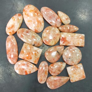 May include: A collection of 15 sunstone cabochons in various shapes and sizes. The stones are a mix of orange, yellow, and red hues with a shimmering, iridescent effect.