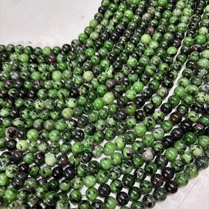 May include: A collection of round beads in shades of green, with black and maroon markings. The beads are strung together, forming a flowing pattern. These beads are suitable for jewellery making.